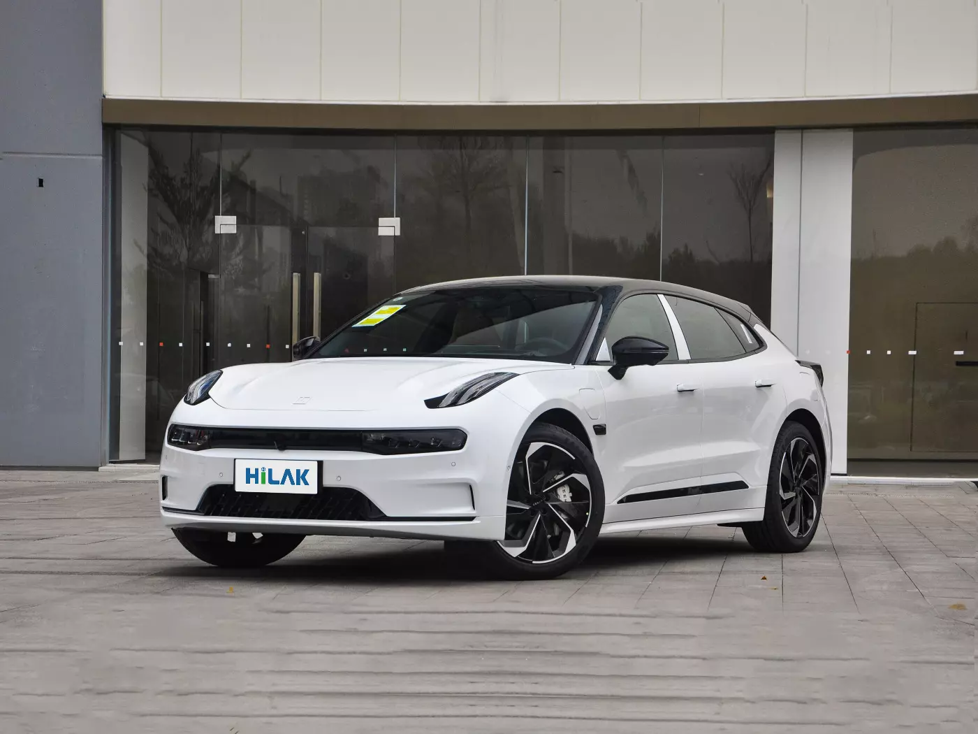 A white ZEEKR 001 electric car is parked on a concrete pavement with a glass building behind it.